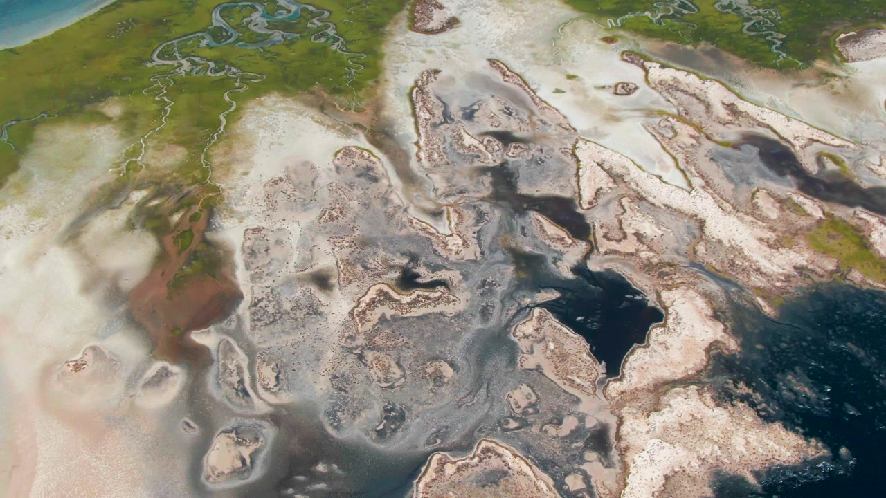 Aerial frame from Guerrero Negro, Baja California Sur, with low fog spreading across pale wetland ground, green marsh channels, and dark blue water, with no visible people.