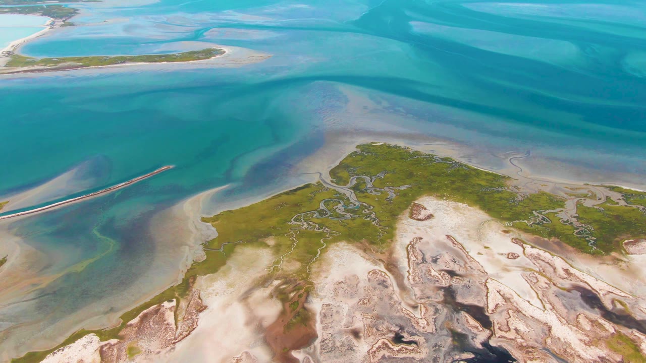 Aerial frame over Guerrero Negro, Baja California Sur showing turquoise lagoon water, pale sandy flats, green island vegetation, a narrow breakwater at left, and no visible people.