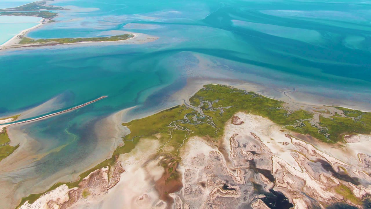 Aerial frame in Guerrero Negro with deeper blue water across the top, pale exposed sand in the lower foreground, green island patches through the center, and no visible people.