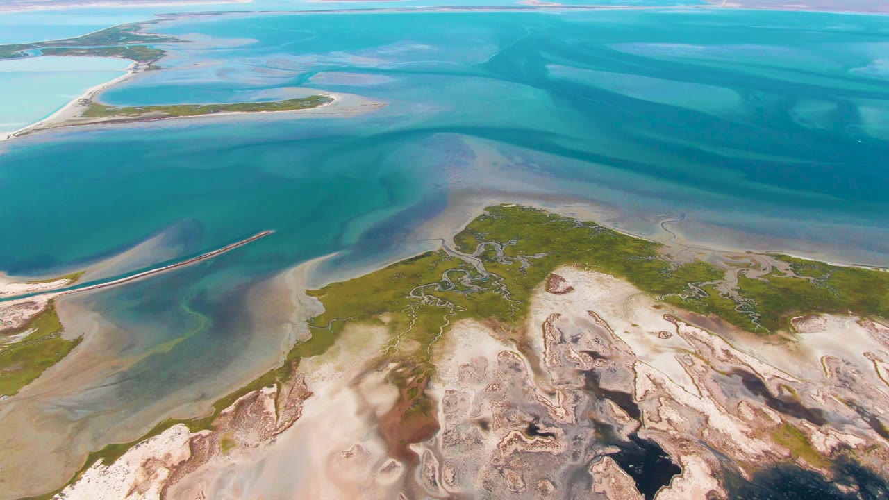 Aerial view over Guerrero Negro tidal water with layered turquoise and blue channels, a small green island near the upper left, sandy shallows below, and no visible people.