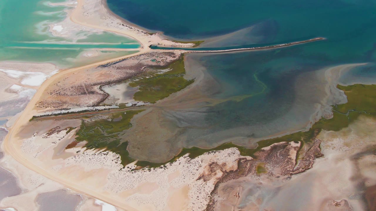 High aerial frame over Guerrero Negro salt flats with beige sand, green marshy patches, shallow blue water, and no visible people.