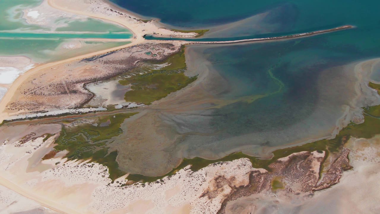 High drone-style frame over Guerrero Negro with turquoise shallows at left, darker lagoon water above, beige flats below, and no visible people.