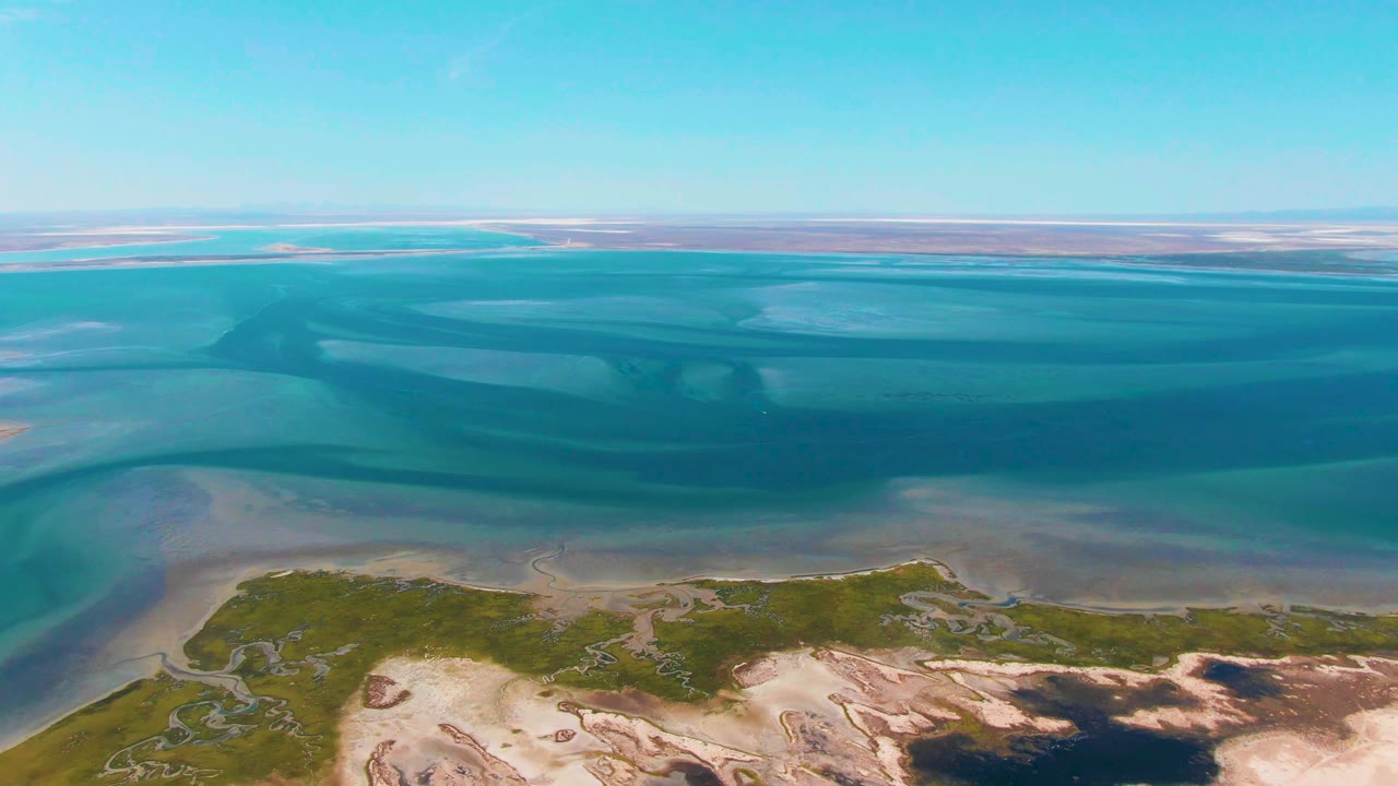 Aerial frame over Guerrero Negro coastal wetland with turquoise water bands, green marsh along pale sand, distant flats on the horizon, and no visible people.