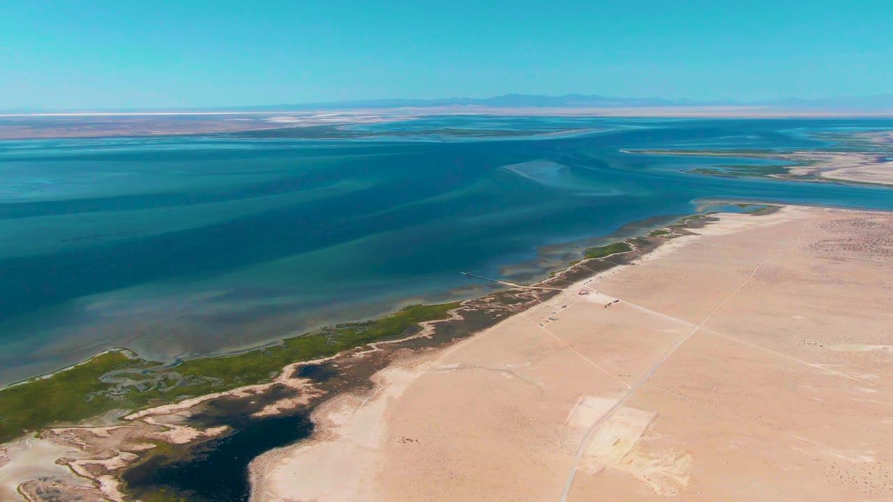 Aerial view of blue coastal water, marsh edge, and pale sandy shoreline.