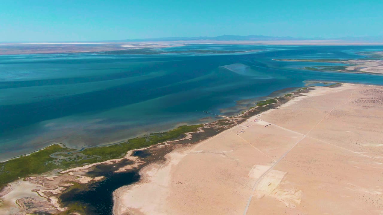 Aerial view in Guerrero Negro with turquoise and deep blue water stretching across the upper frame, green marsh in the middle, pale sand below, and no visible people.