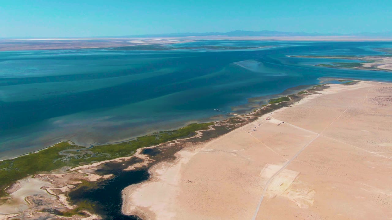 Oblique aerial frame over Guerrero Negro showing the waterline curving beside green marsh patches and wide pale sandy ground, with no visible people.