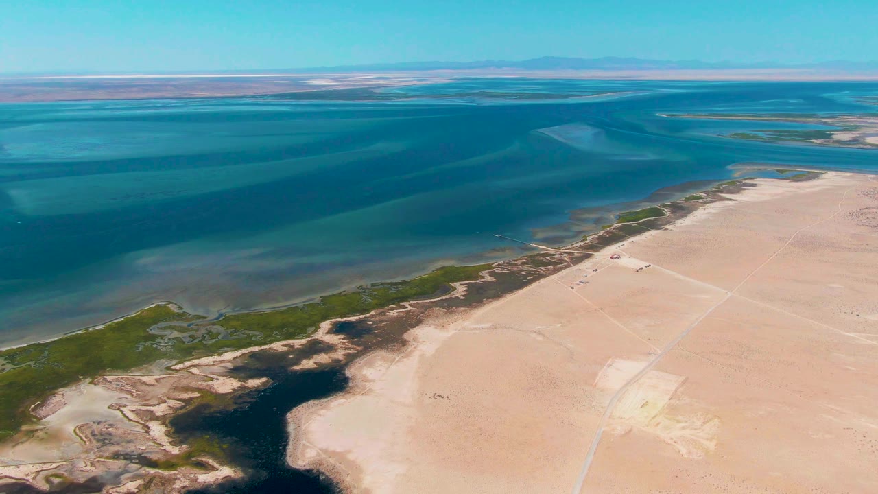 Aerial frame in Guerrero Negro with broad blue water occupying the upper view, a green marsh strip along the shoreline, pale sand below, and no visible people.