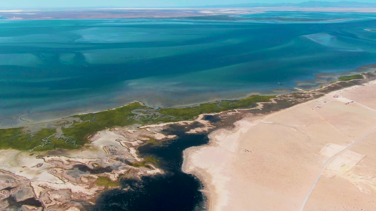 Opening aerial frame over Guerrero Negro, Baja California Sur, with turquoise coastal water above green wetlands and dark inlet water beside beige sand, no visible people.