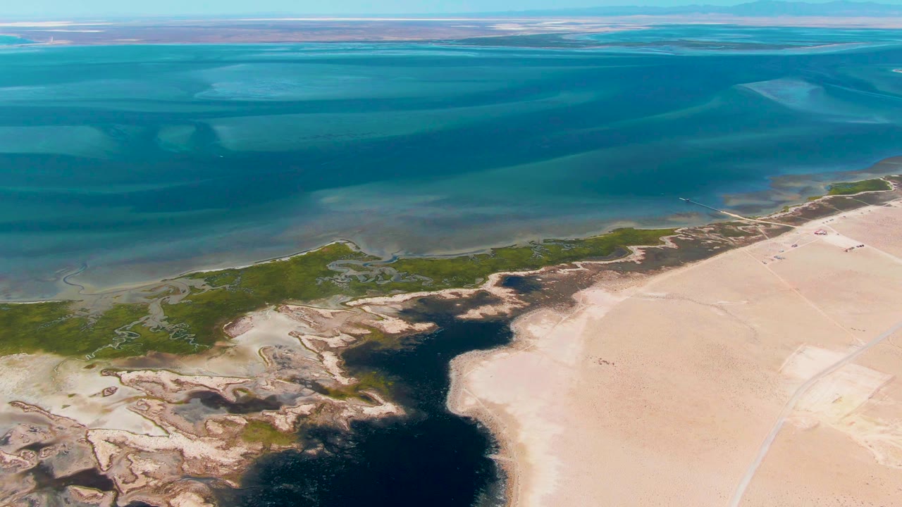 Aerial frame from Guerrero Negro showing the dark inlet extending inland between green marshy patches and light sand flats, with open blue water beyond and no visible people.