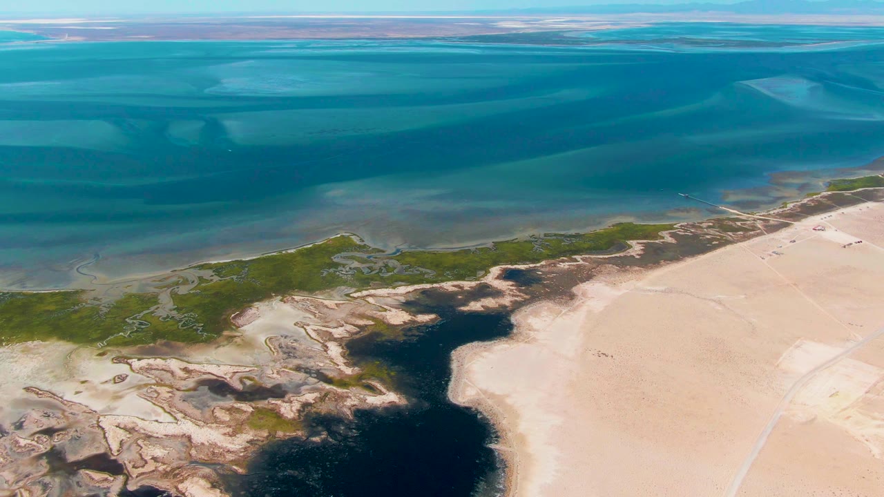 Mid-clip aerial view in Guerrero Negro with layered blue ocean in the background, green coastal wetlands along the shore, and beige sand flats around a dark tidal pool, no visible people.