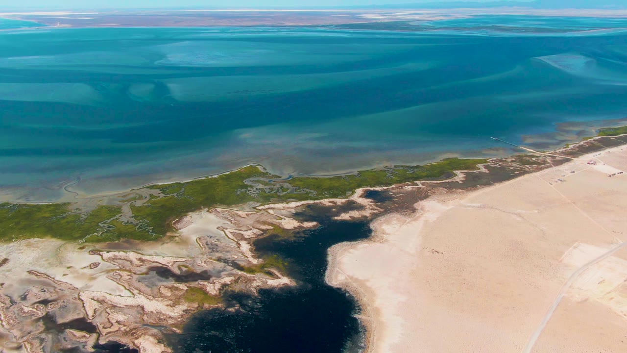 Aerial coastal inlet with shallow blue water, wetland greens, and pale sand.