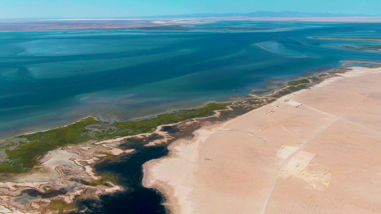 Aerial frame over Guerrero Negro, Baja California Sur, with blue shallow water across the top, pale sand at lower right, green vegetation at lower left, dark channel water near shore, and no visible people.