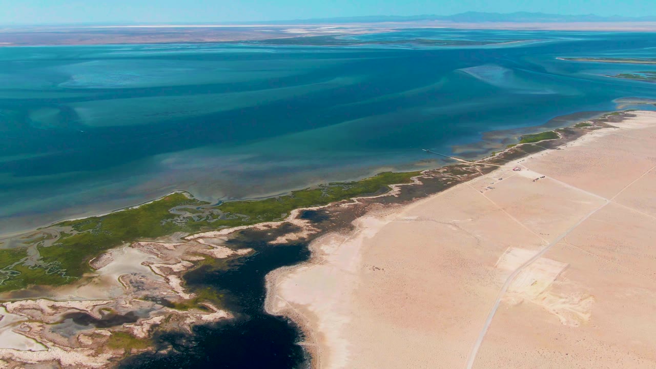 Drone-style aerial frame over Guerrero Negro, Baja California Sur, with shallow blue water above a sandy coast, dark pooled water near green vegetation, a pale track in the sand, and no visible people.