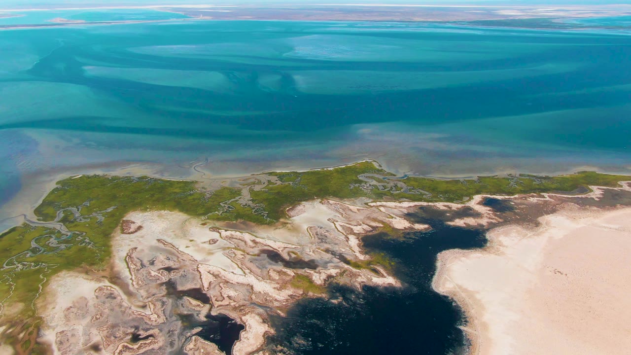 Mid-clip aerial frame in Guerrero Negro showing turquoise coastal water above green vegetation, pale sand flats, and dark wet channels, with no visible people.