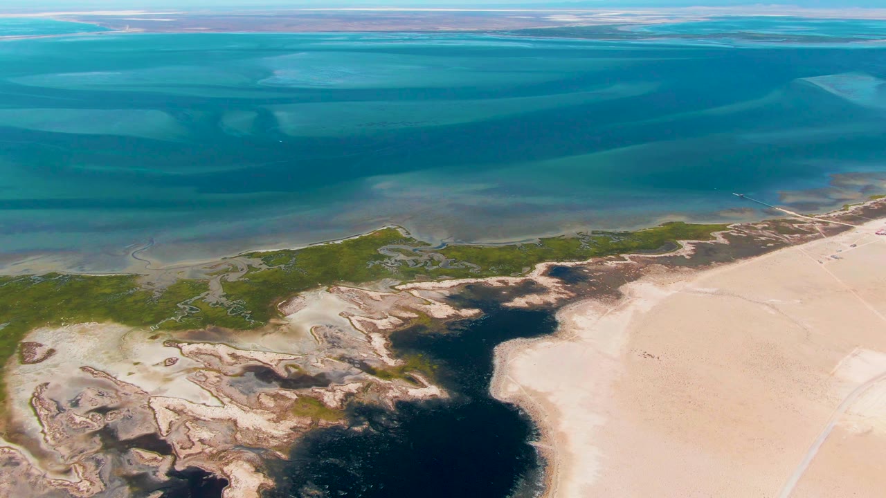 Aerial frame over Guerrero Negro, Baja California Sur showing turquoise ocean bands above green marsh, dark wet channels, pale sand, and no visible people.