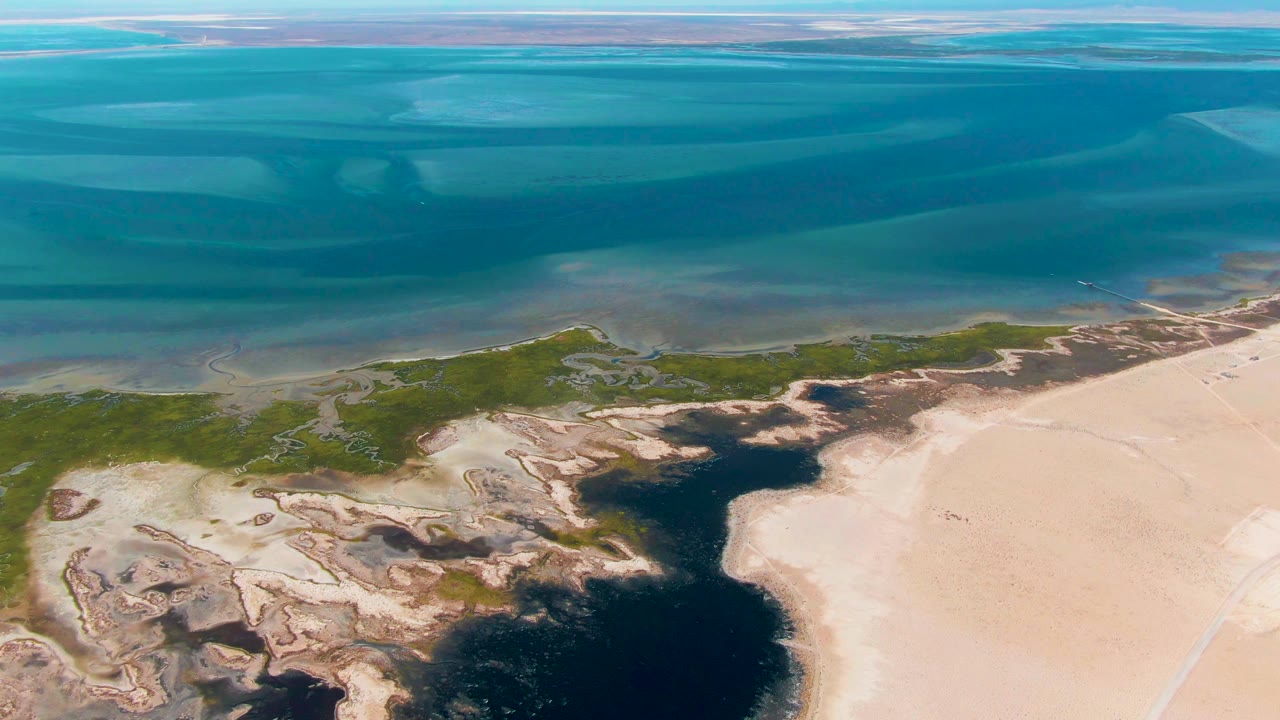 Aerial view at 0:01 over Guerrero Negro with layered blue water, a narrow marsh strip, pale beach sand, dark tidal pools, and no visible people.