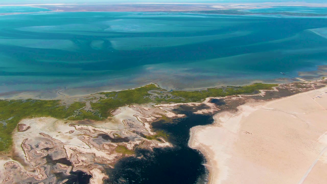 Frame 0:02 shows Guerrero Negro coastline from above, with shallow turquoise water meeting green marsh patches, dark wet inlets, pale sand, and no visible people.