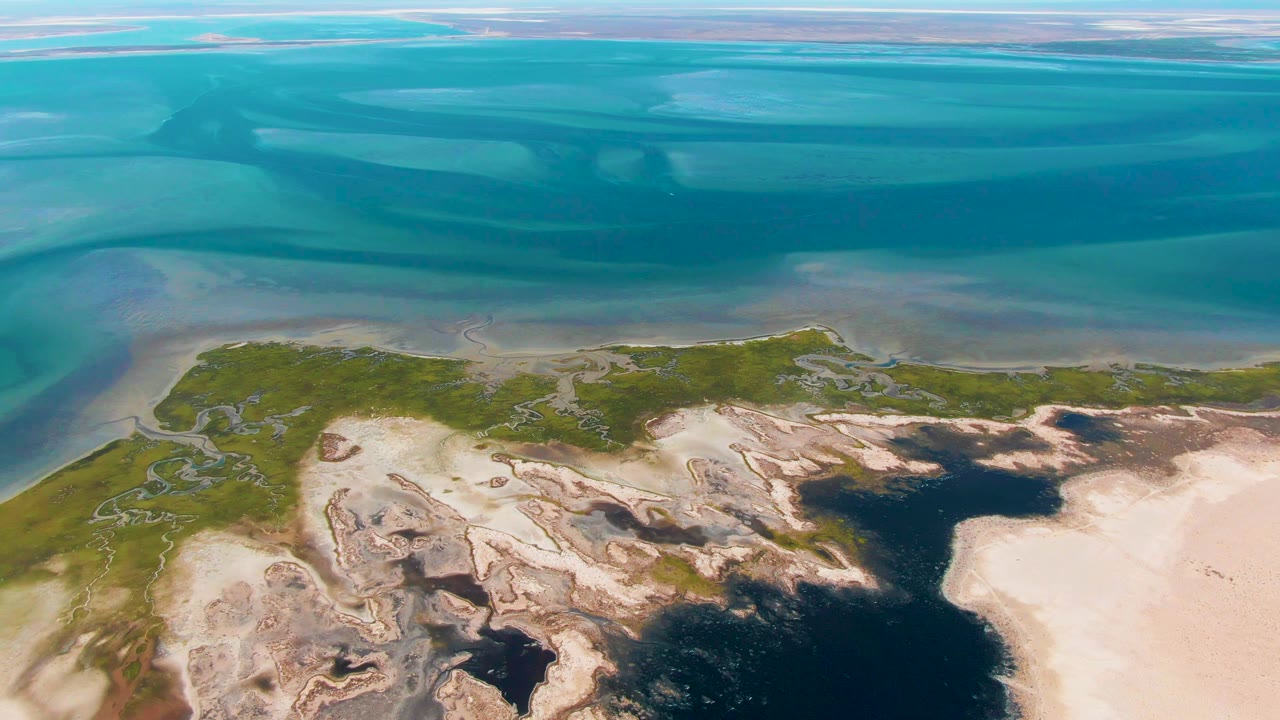 Aerial view in Guerrero Negro with shallow blue water across the top, sand flats and rocky textures below, green patches near the shore, and no visible people.
