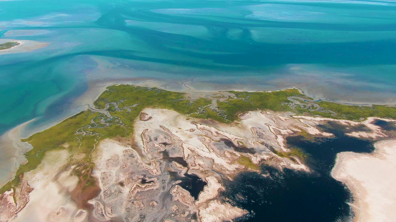 High aerial view in Guerrero Negro, Baja California Sur, where layered blue water sits above pale sand flats and green shoreline, no visible people.