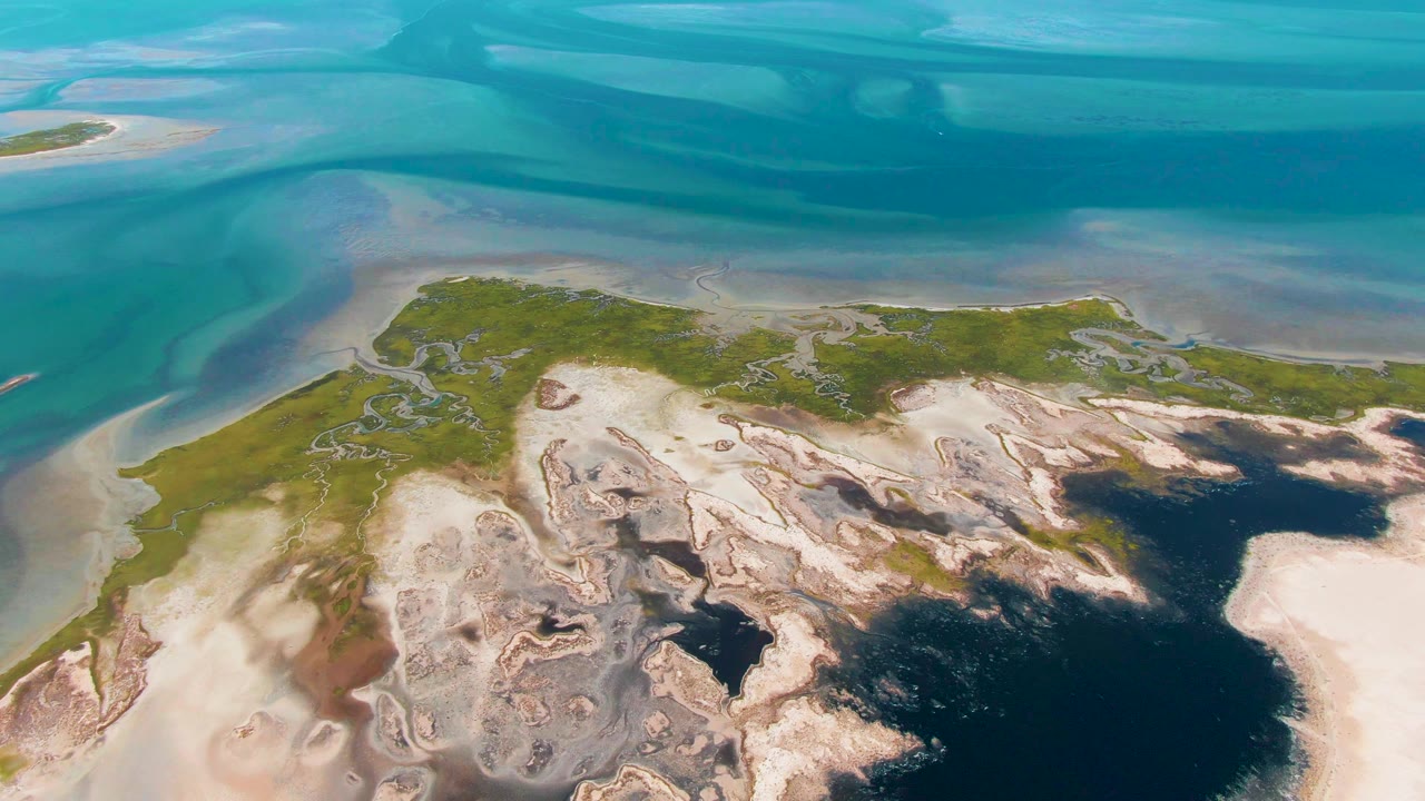 Mid-clip aerial view over Guerrero Negro, Baja California Sur, with blue coastal water across the top and pale sand flats around a green landmass, no visible people.
