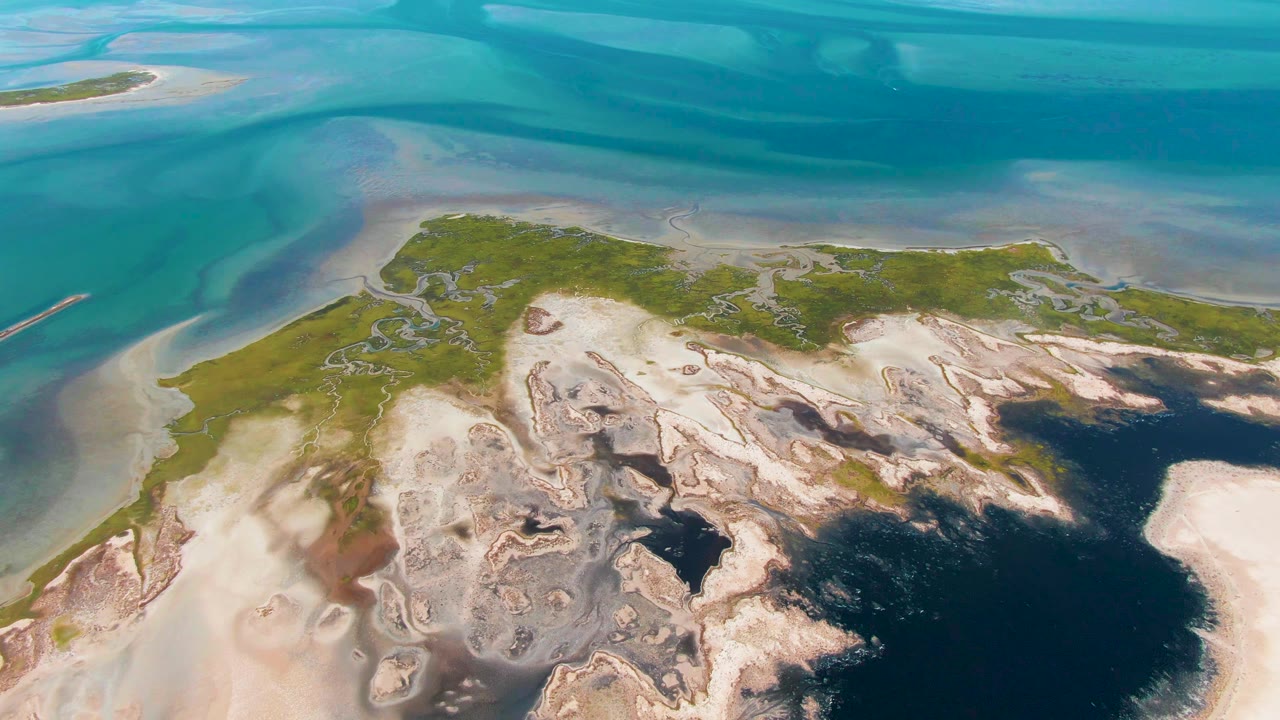 Opening aerial frame over Guerrero Negro tidal flats, with turquoise shallow water above pale exposed sand, gray mud patterns, dark pools, green vegetation, and no visible people.