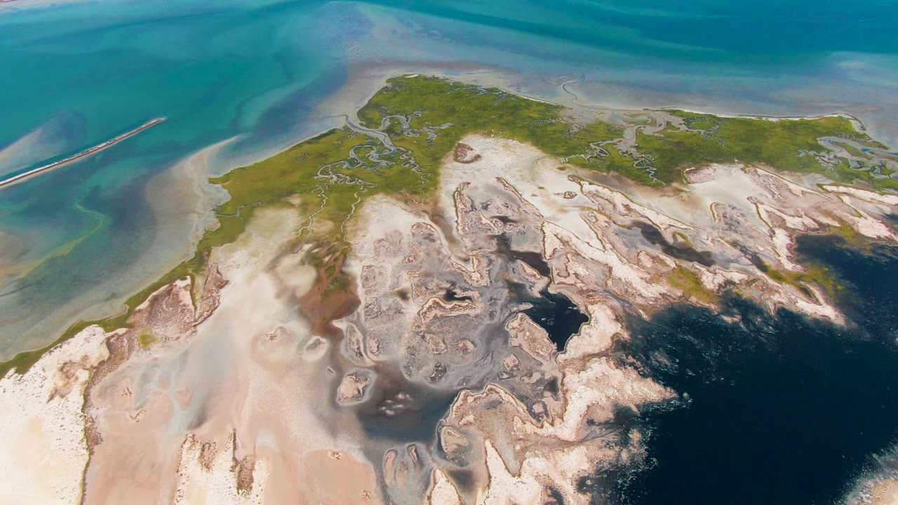 Aerial view over Guerrero Negro with braided pale sand patterns, patches of green vegetation, shallow turquoise water, a dark lower-right shadow, and no visible people.