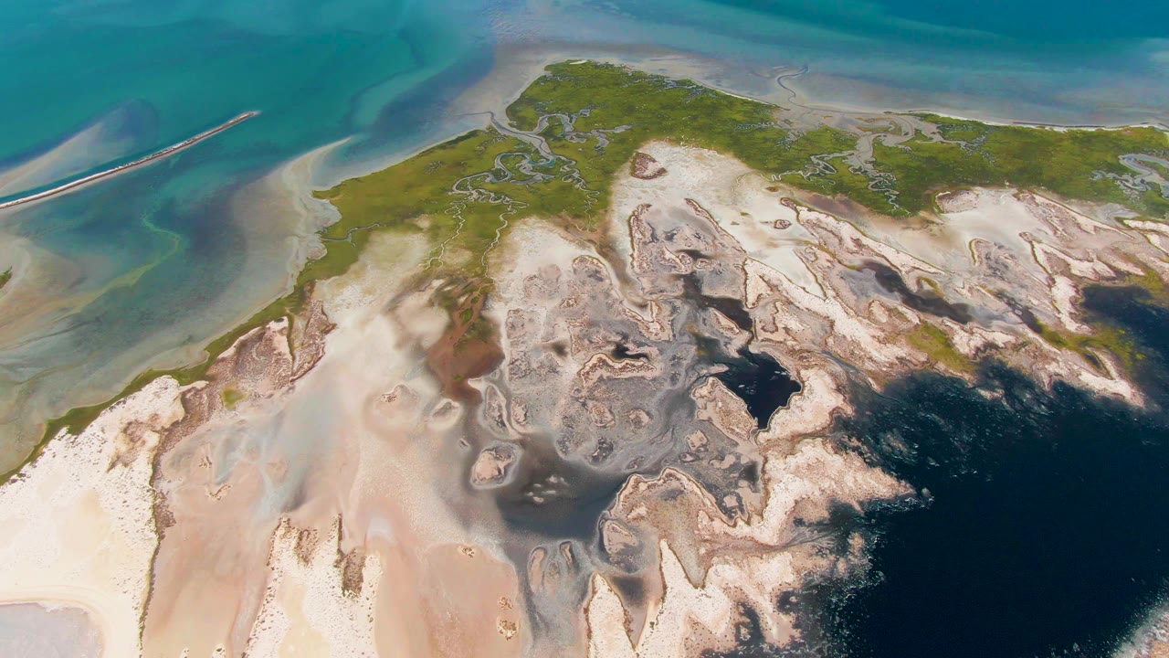High aerial view over Guerrero Negro coastal sand flats, green land, turquoise shallows, and darker water, with no visible people.