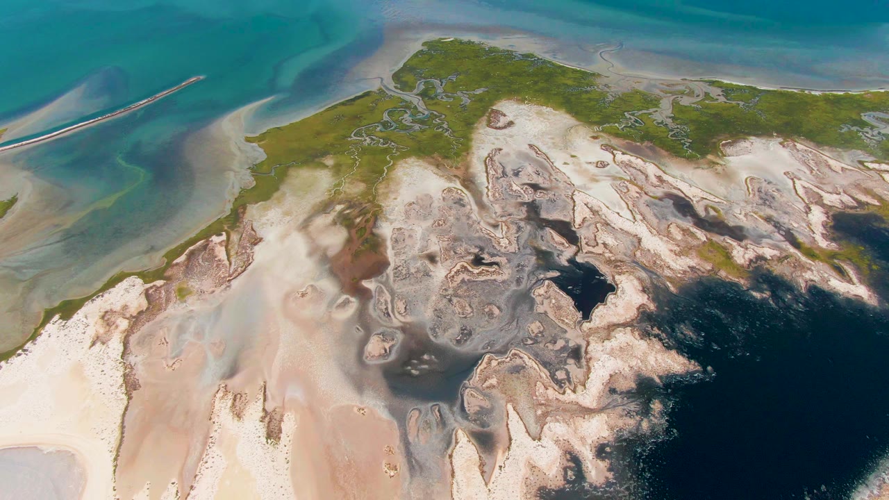 Drone-like overhead frame in Guerrero Negro showing green land at the upper center, pale sand and mud flats below, and blue-green water along the top, with no visible people.