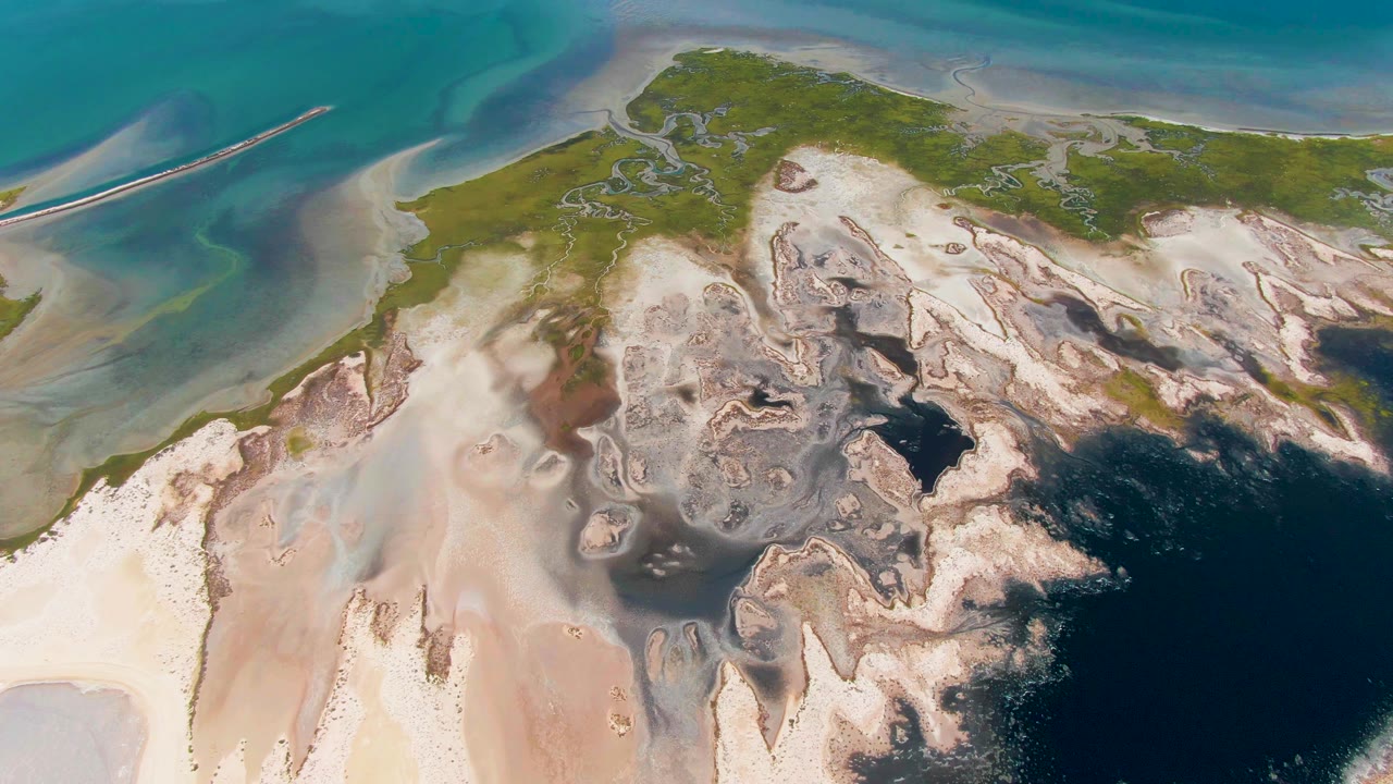 Overhead Guerrero Negro coastal frame with mottled mud flats, white sand, green shoreline, turquoise shallows, and a darker water area at lower right, with no visible people.