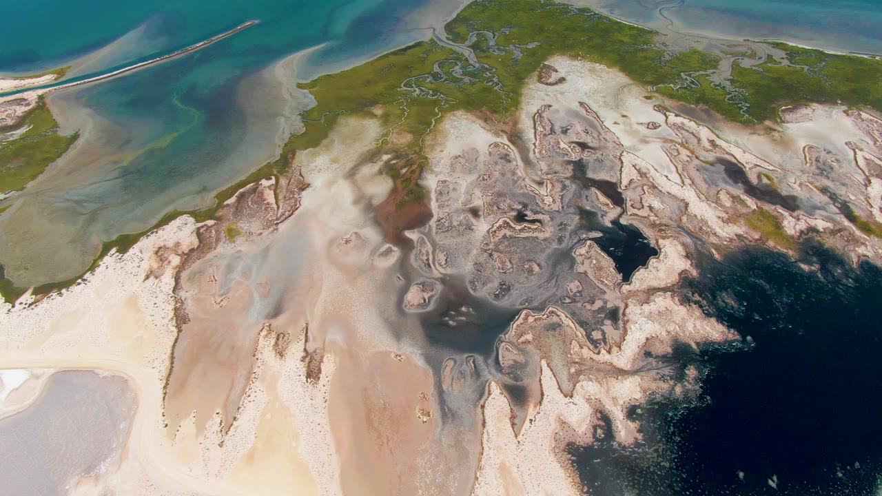 A high aerial view over Guerrero Negro coastal flats shows pale shoreline, green vegetation, dark blue water, and a small boat beginning its left-to-right path near the upper left, with no visible people.
