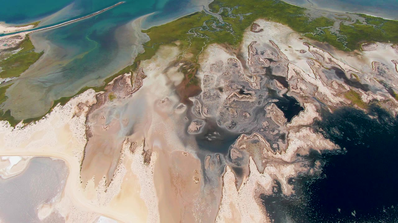At 0:02, the Guerrero Negro coast appears as layered green, white, brown, and deep blue shapes, with the small boat continuing rightward near the top of frame and no visible people.