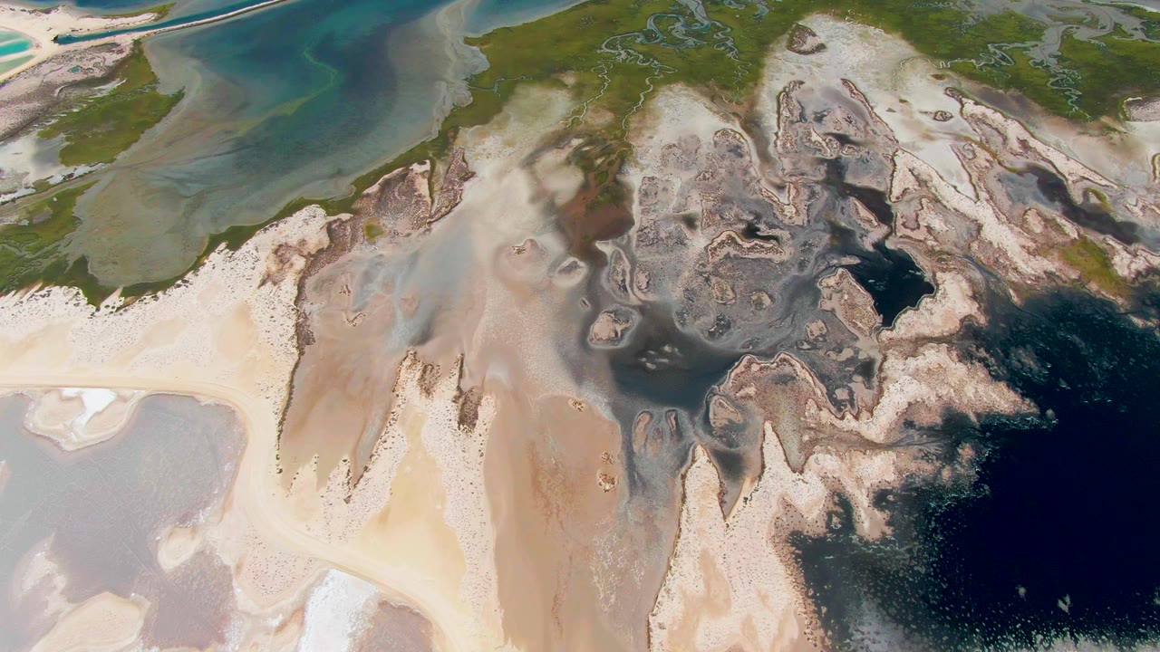 Top-down aerial view in Guerrero Negro with beige mud flats, pale sand channels, green marsh edge, and a dark water pocket at the right, no visible people.