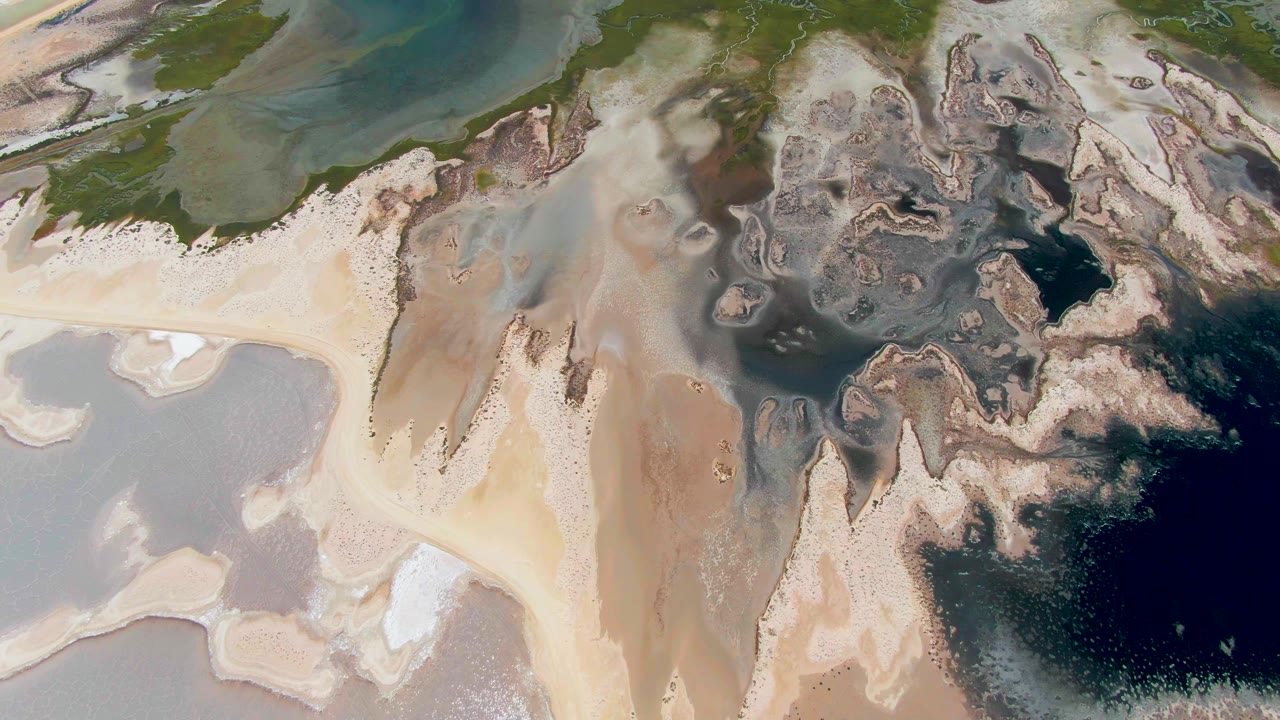 Overhead view of Guerrero Negro tidal flats with pale sandbars, gray-green shallows, dark water pockets, mottled sediment, and no visible people.