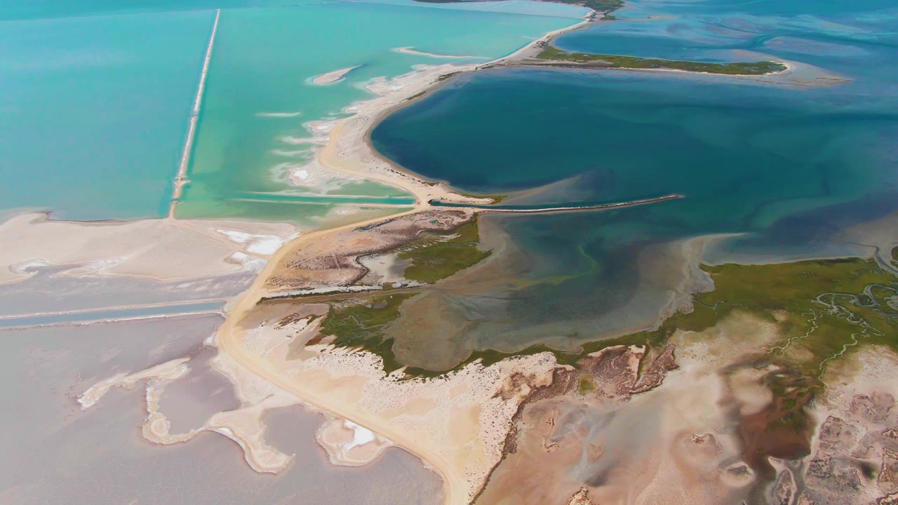 Aerial view of sandy coastal road, turquoise water, salt flats, and green shore patches.