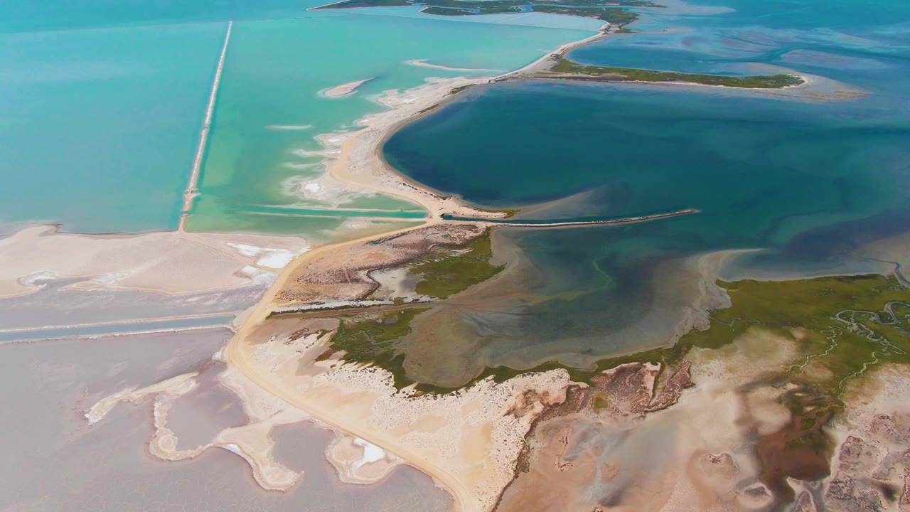 At 0:02, Guerrero Negro's coastal road and shore remain in high oblique view, with the causeway crossing bright turquoise water above darker shallows and no visible people.
