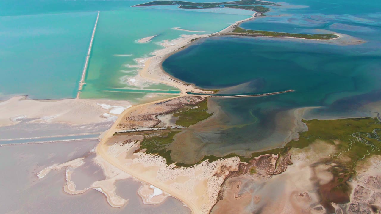 At 0:03, the aerial view shifts slightly along the Guerrero Negro shoreline, with pale sandbars at lower left, a road crossing the frame, green vegetation below, and no visible people.