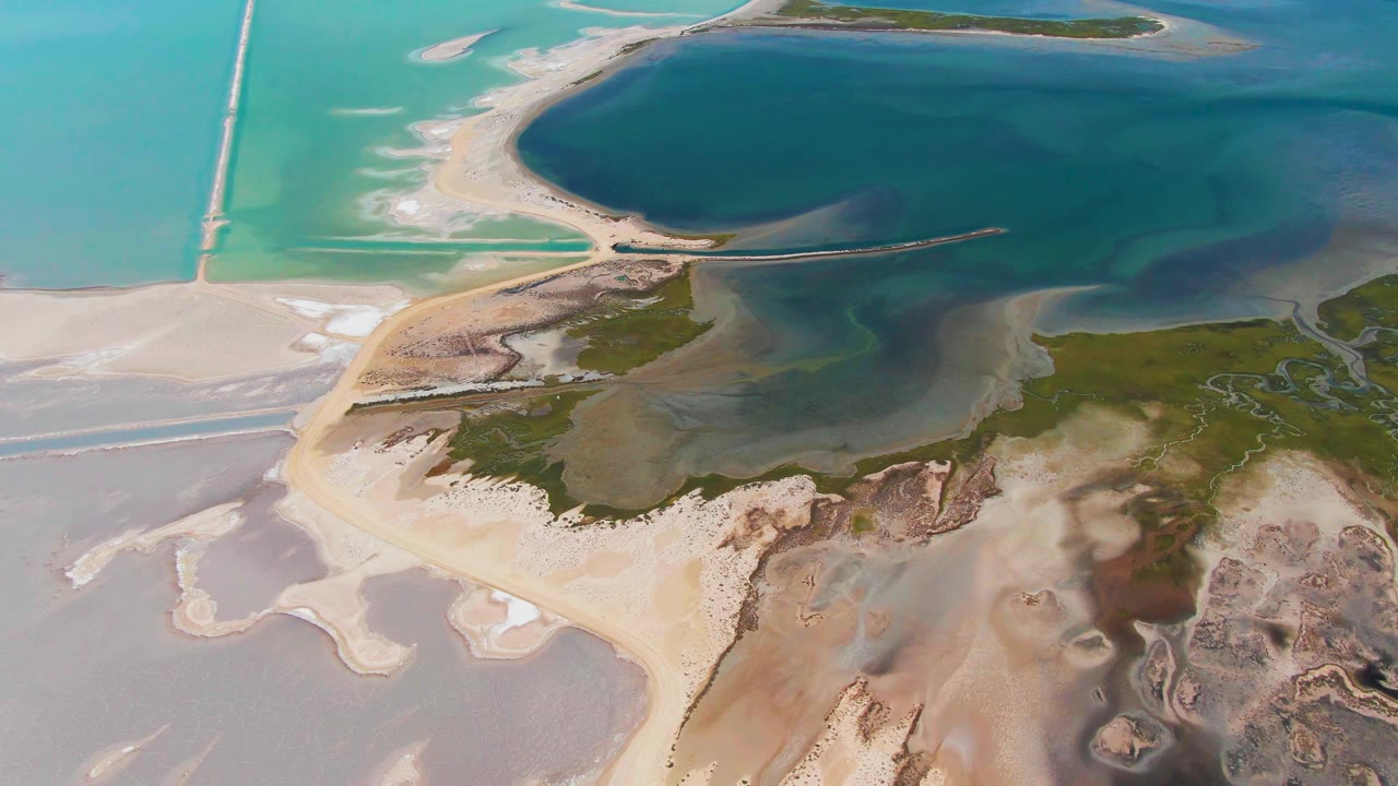 Aerial view over Guerrero Negro, Baja California Sur, Mexico, showing a pale sandbar beside turquoise and dark blue lagoon water, a straight causeway, marshy green patches, and no visible people.