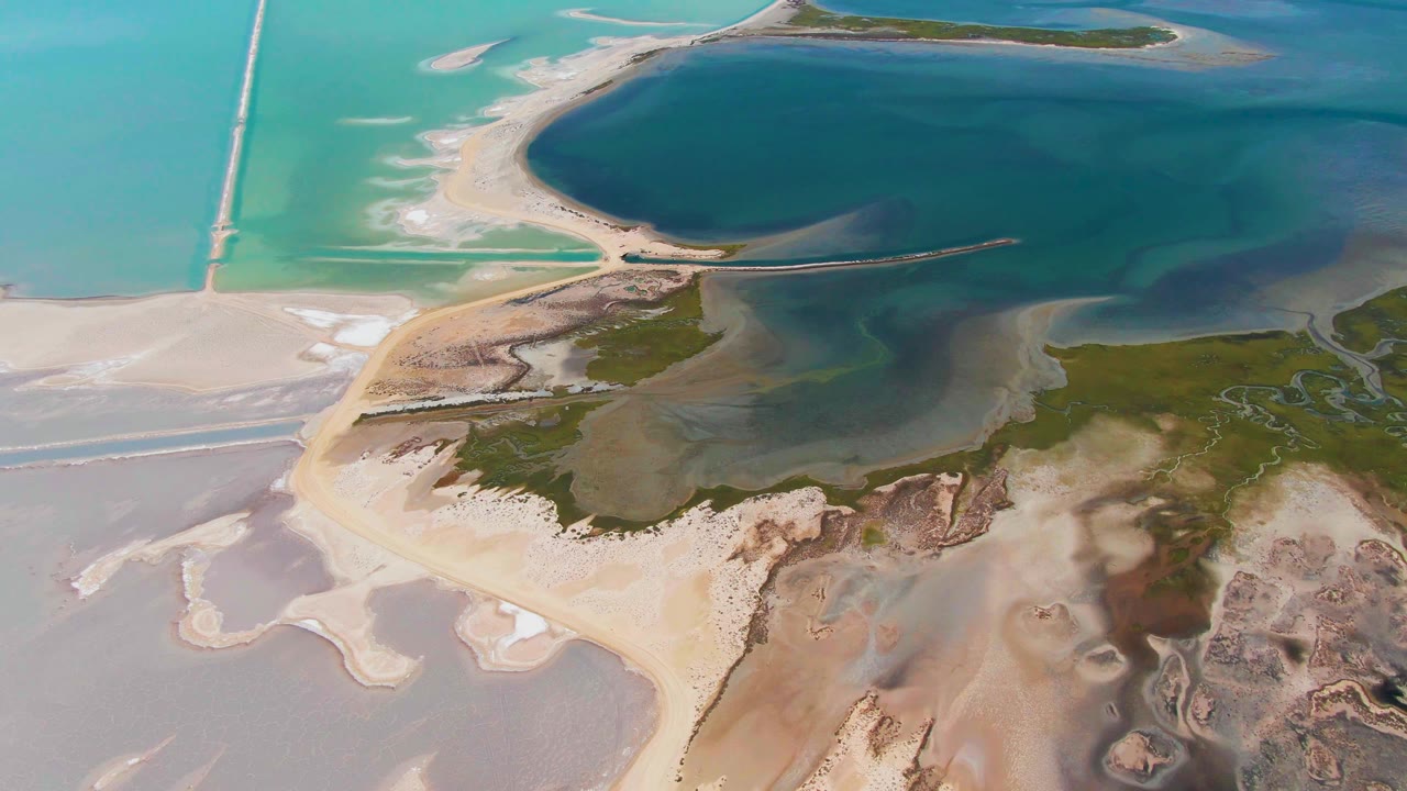 Aerial frame from Guerrero Negro with the causeway near the top, deep blue lagoon water to the right, pale sand flats to the left, green marsh texture below, and no visible people.