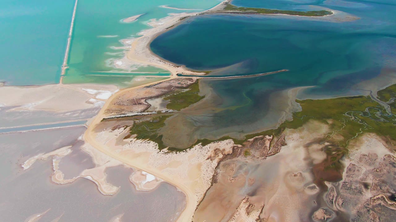 High aerial view in Guerrero Negro, Baja California Sur, Mexico, with a curved sandy shoreline, dark lagoon water, turquoise shallows, small channel marks in green marsh, and no visible people.