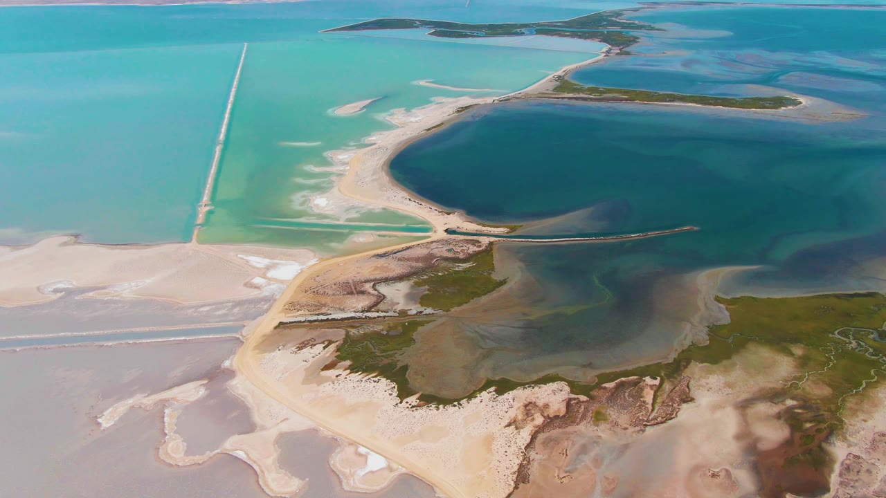 Aerial frame over Guerrero Negro, Baja California Sur showing turquoise shallows, a dark blue pool, pale salt flats, a long causeway, and no visible people.