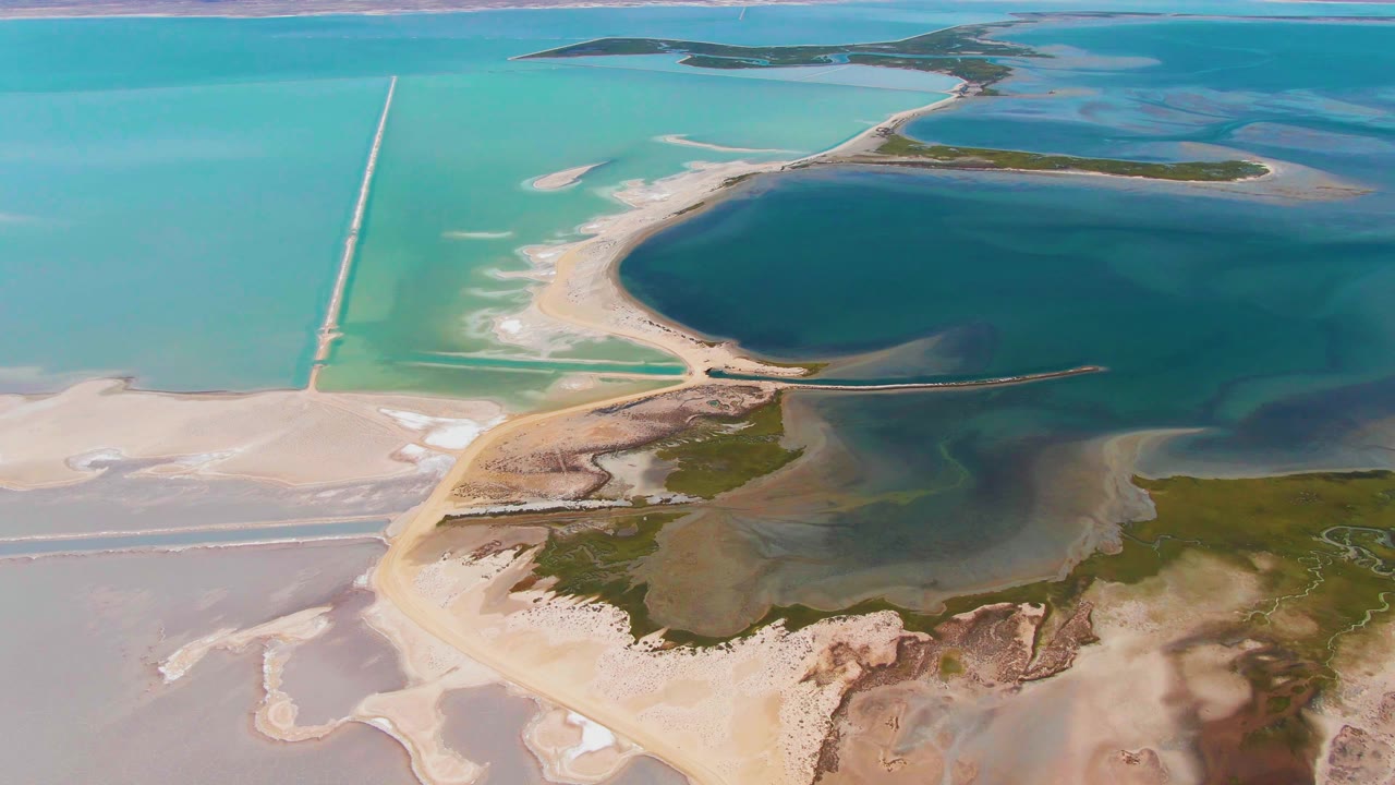 Aerial frame over Guerrero Negro with a dark blue lagoon bordered by pale sand, salt-encrusted flats, green patches, road lines, and no visible people.