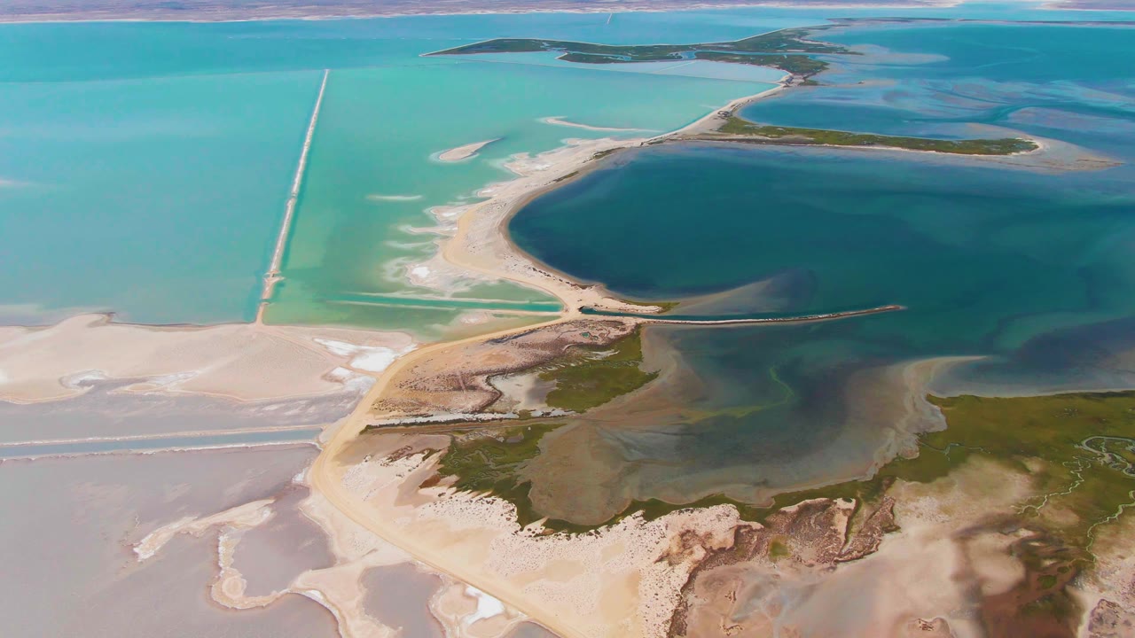 Aerial frame in Guerrero Negro showing turquoise shallows crossed by a straight causeway, a rounded dark pool, sandbars, distant islands, and no visible people.