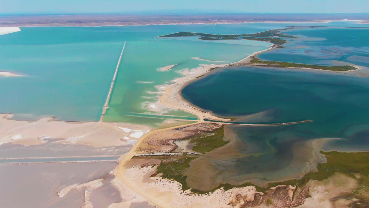 Aerial frame in Guerrero Negro with the causeway spanning pale turquoise shallows, a curved sandy point below it, dark blue-green lagoon water to the right, distant islands, and no visible people.