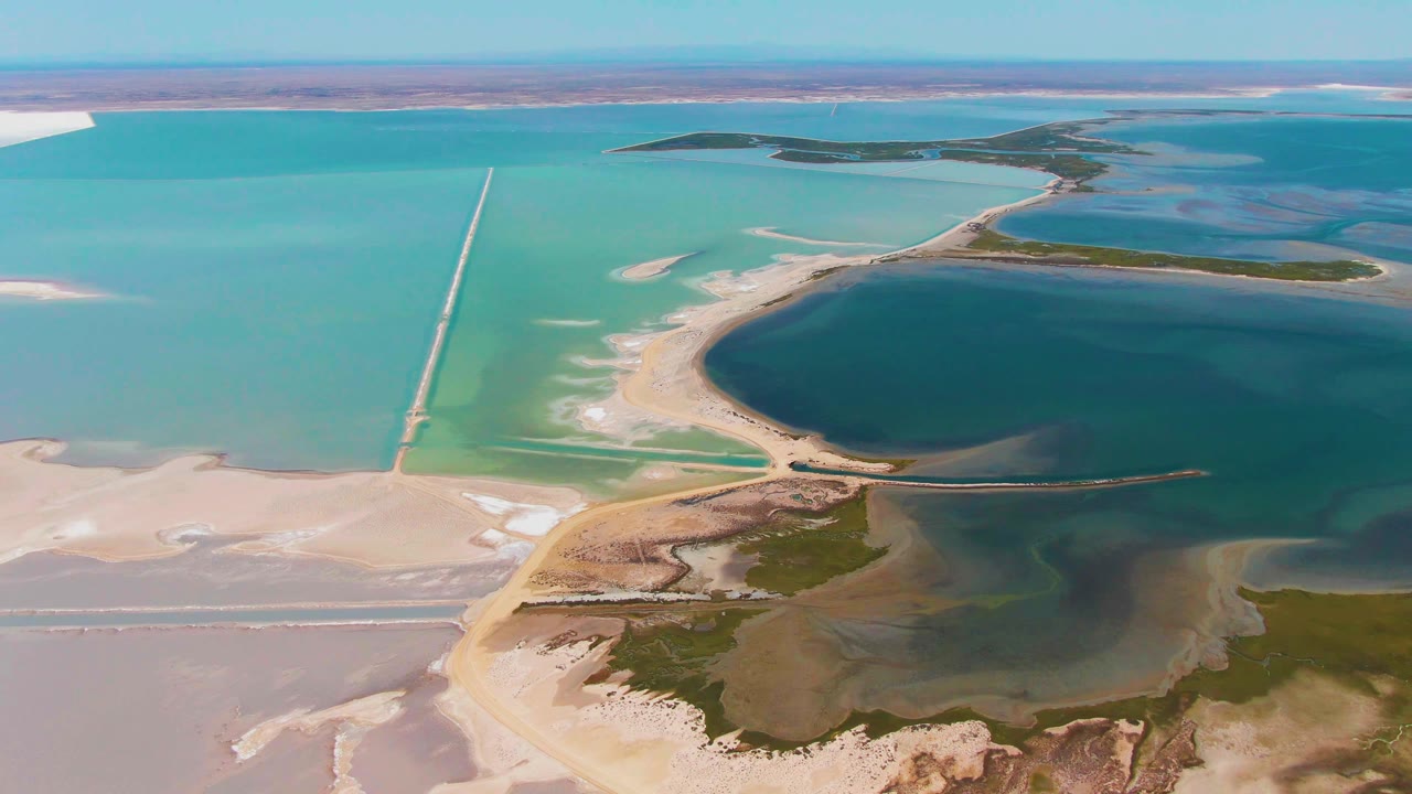 Mid-clip aerial view over Guerrero Negro shows the causeway running through turquoise water, broad exposed salt flats in the lower left, green vegetation near the darker lagoon edge, and no visible people.