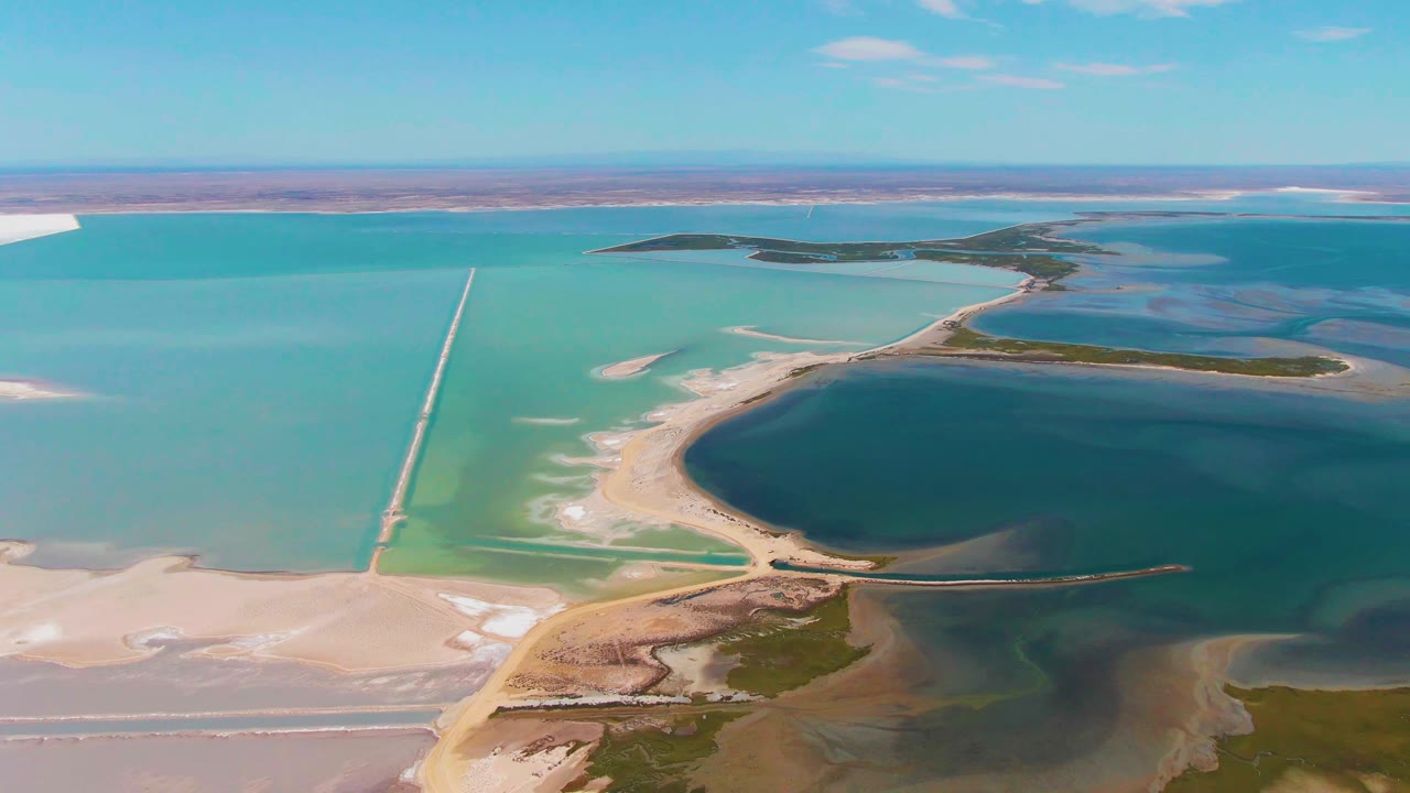 Early aerial frame over Guerrero Negro salt lake water, with a straight pale causeway at left, a curved sandbar around deep blue water, low islands beyond, and no visible people.
