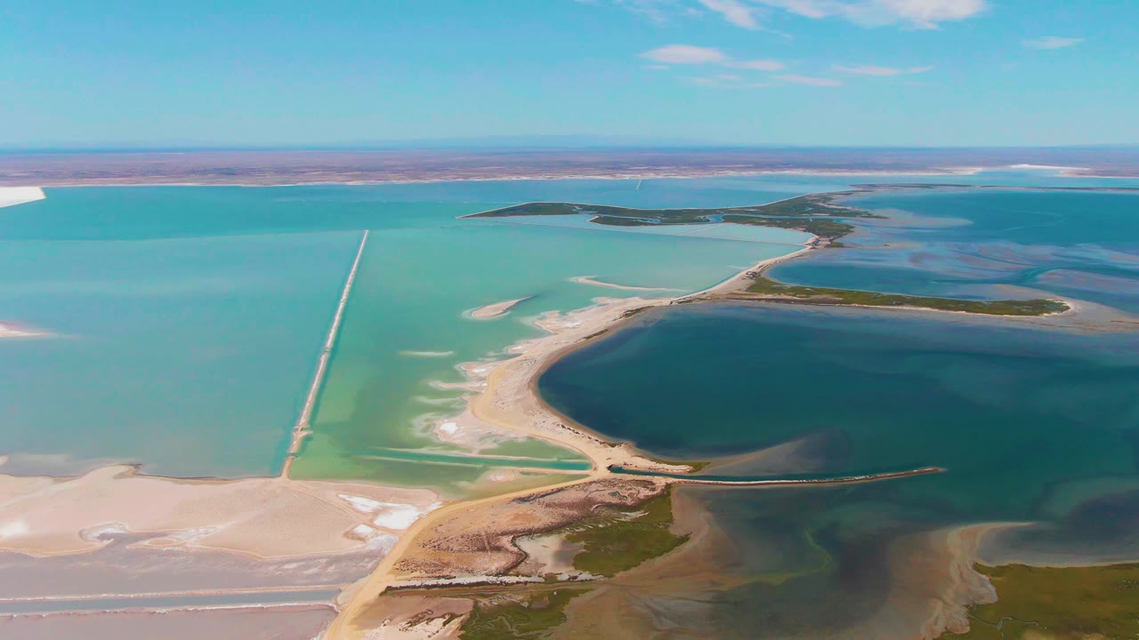 At 0:02, the Guerrero Negro salt lake remains in a wide aerial composition, showing the long causeway beside a pale curved shore, greenish islands, layered water colors, and no visible people.
