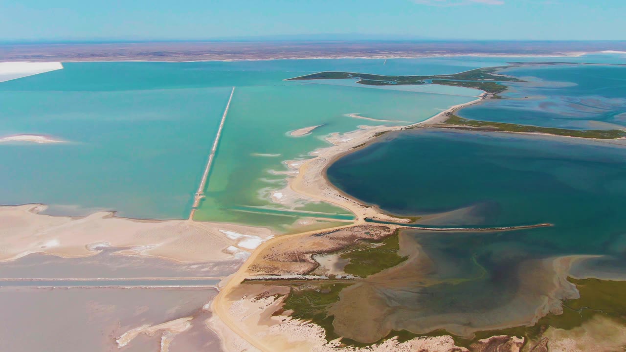 Aerial view over Guerrero Negro salt evaporation ponds with a straight causeway crossing blue-green water, pale salt flats, dark lagoon water, and no visible people.