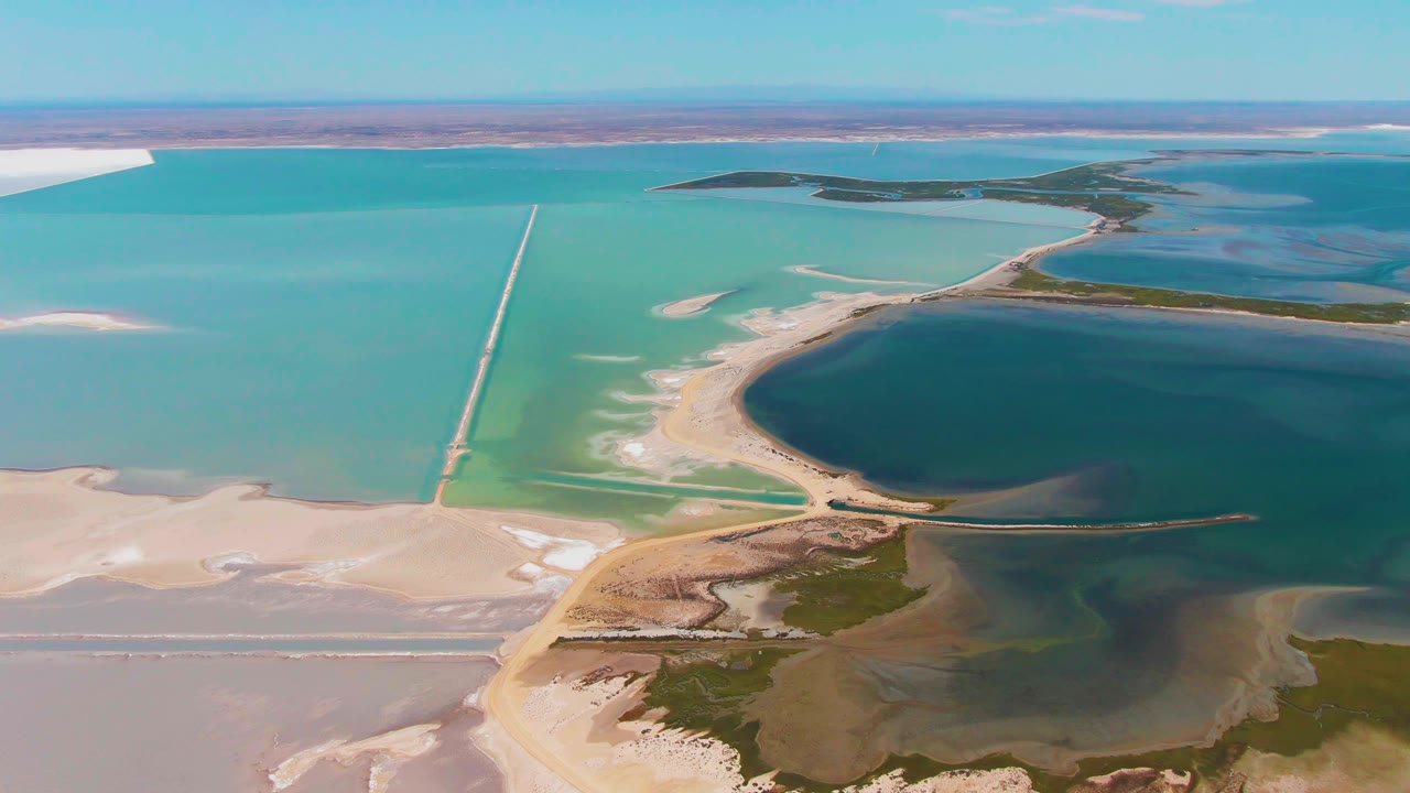 High aerial frame in Guerrero Negro showing turquoise evaporation ponds, a dark rounded water basin, pale salt-encrusted shoreline, a long causeway, and no visible people.