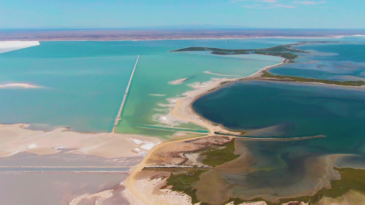 Guerrero Negro aerial frame with the causeway running through blue-green salt ponds toward distant lagoon water, exposed pale flats, and no visible people.
