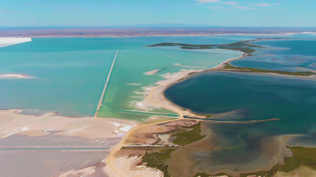 Wide aerial view in Guerrero Negro with salt flats in the foreground, a narrow causeway at left center, green-blue evaporation water, dark lagoon water, and no visible people.